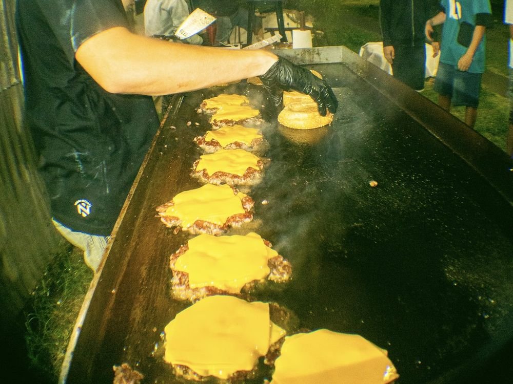 Plancha llena de hamburguesas con cheddar durante un evento de noche, con invitados alrededor